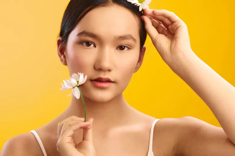 asian woman holds a flower close to her face in a warm studio setting, calm confident expression, soft lighting, minimalist background, creating a serene and elegant portrait.