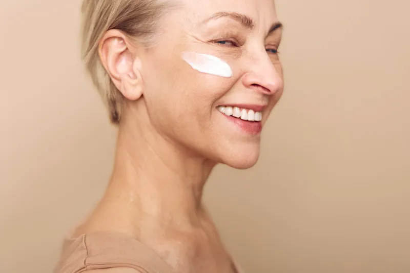 Smiling mature woman with short hair applies white facial cream on cheek. Close-up on beige background. Concept of skincare, aging skin care, wellness, beauty routine, and self-care.