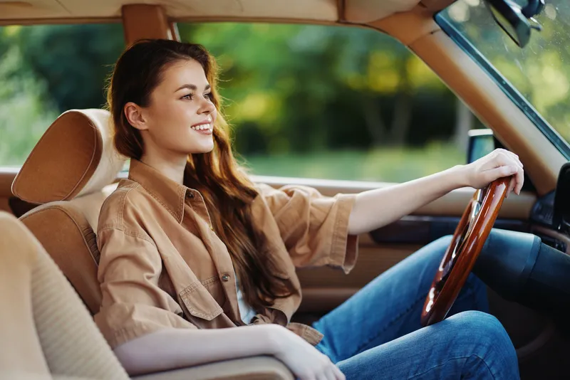 Young woman driving a vintage car, smiling joyfully, wearing a casual shirt and jeans, surrounded by a lush green background, capturing a sense of freedom and adventure.