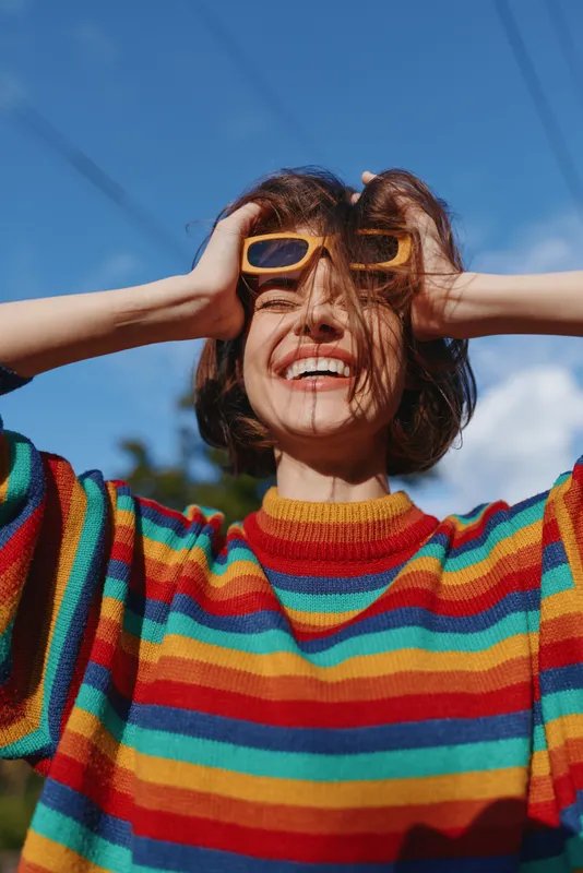Woman young adult smiling in a colorful striped sweater and yellow sunglasses, hands in hair outdoors under blue sky. Casual style, joyful lifestyle portrait for fashion and travel.