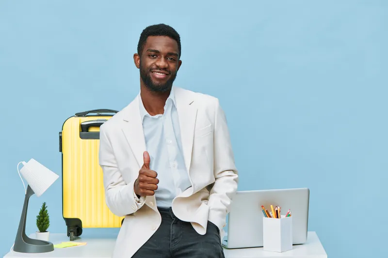Confident smiling man with African descent in stylish white blazer and light blue shirt standing at the modern office desk, giving thumbs up gesture, bright yellow suitcase, laptop, pen holder, minima