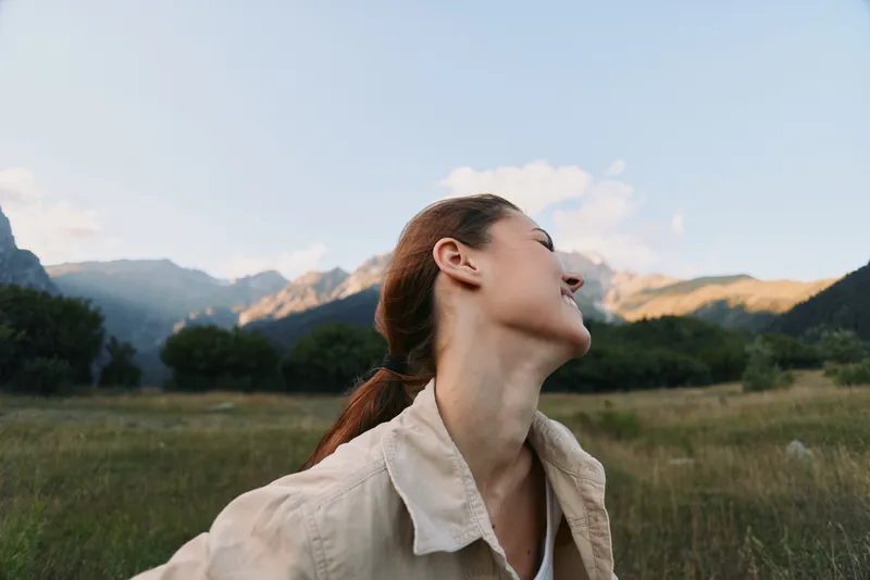 Happy woman outdoors in nature, smiling with eyes closed, enjoying fresh air, casual clothing, green meadow and mountains background, peaceful moment