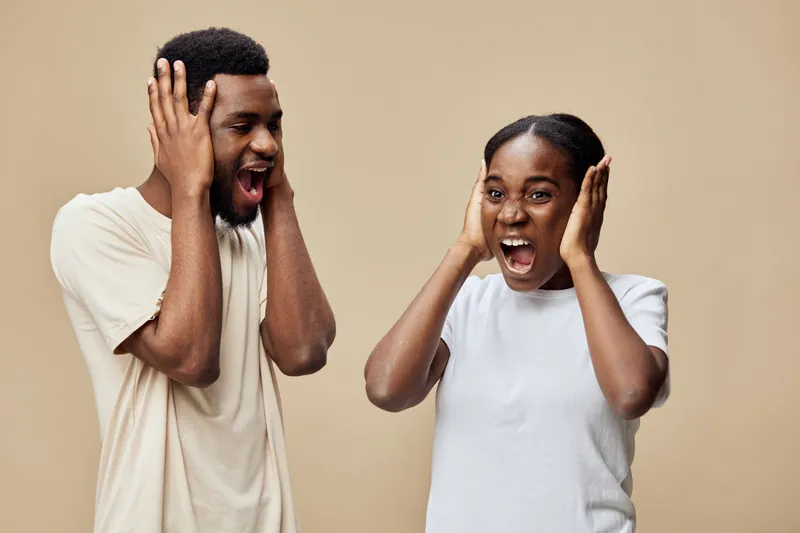 Excited young couple with joyful expressions covering their ears against a soft beige background, showcasing happiness and energy in a modern style