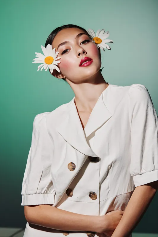 Fashionable young woman with bold red lips and two white daisies in her hair poses confidently against a soft green background. Creative makeup, beauty theme.