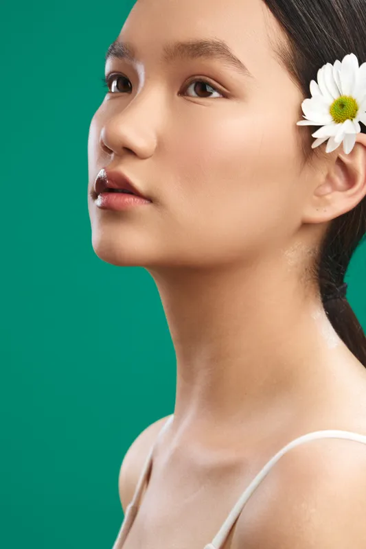 Asian woman with a delicate flower tucked behind her ear gazes softly off camera, captured in a studio portrait with a vibrant green backdrop, conveying calm beauty and graceful poise.