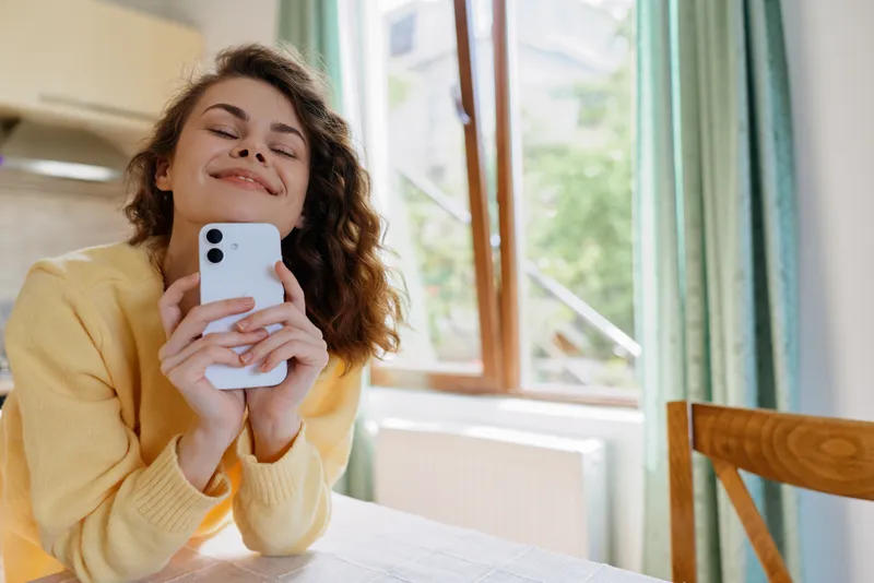 Smiling woman with curly hair holding a smartphone indoors at a sunlit kitchen table. Warm natural light, relaxed home life, casual beauty, wellness, and self-care mood.