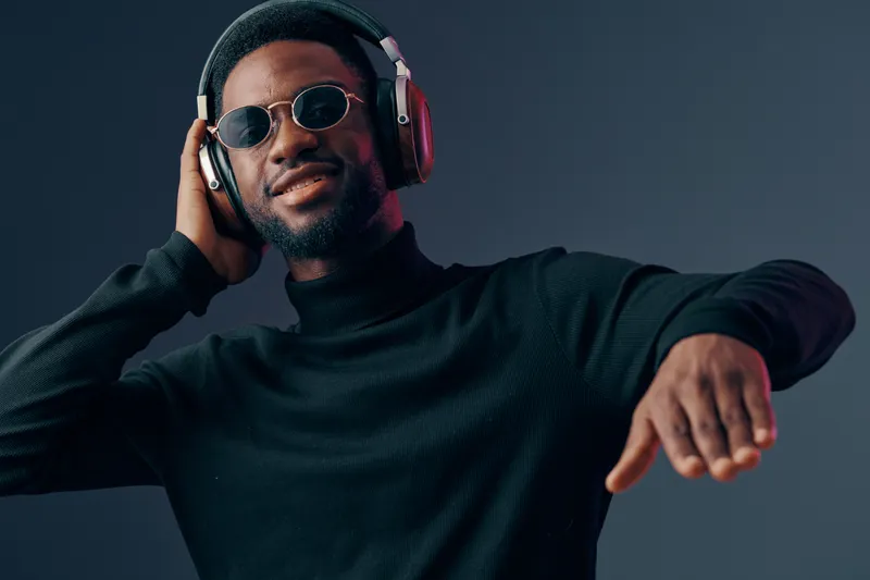 Young Black man enjoying music with headphones, smiling confidently in a stylish black outfit under soft lighting, creating a vibrant and modern atmosphere