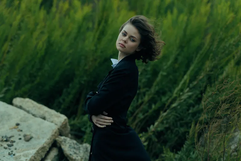 Woman in a dark coat stands with arms crossed on rocky shore, leaning back against a green forest, captured in moody outdoor setting with wind blowing hair