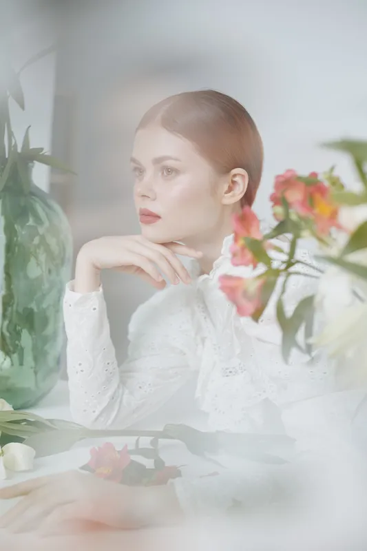 Elegant young woman in white blouse, surrounded by fresh flowers and greenery, exuding calmness and beauty in a soft, natural setting
