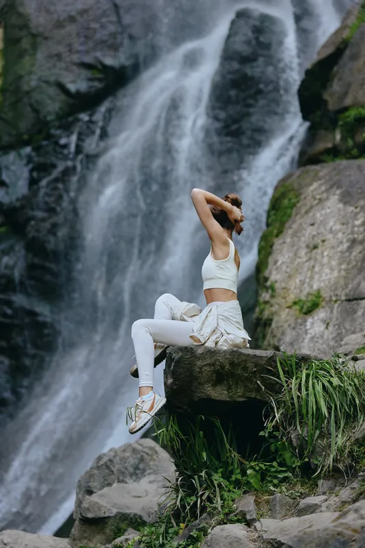 woman relaxing by waterfall in white activewear sitting on stone with green foliage around, calm natural outdoor scene showcasing peaceful meditation and nature connection