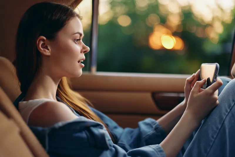 Young woman enjoying a mobile game in a car at sunset, showcasing emotions of excitement and joy, dressed casually in denim with a natural backdrop.