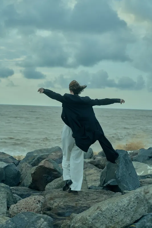 Woman with outstretched arms stands on rocky shore, facing the sea under cloudy sky, wind-swept coat and white trousers, conveying freedom, balance and vast ocean mood