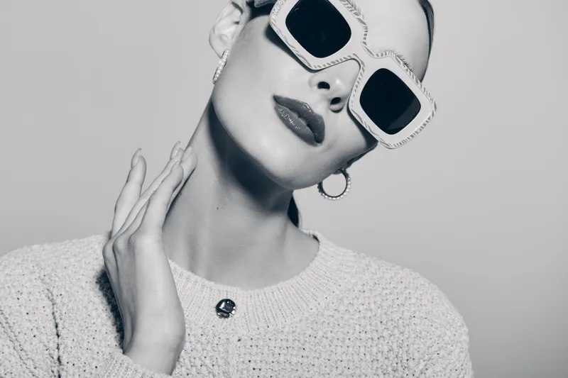 Stylish white woman with fair skin wearing large sunglasses and hoop earrings poses in knitted sweater against neutral background. Concept for fashion, elegance, and modern style.