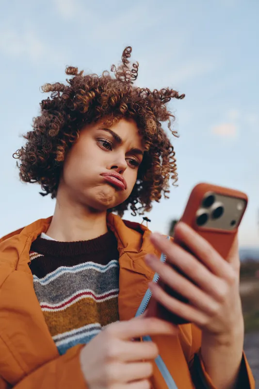 Woman lifestyle portrait, curly hair young woman outdoors, holding smartphone selfie, orange jacket, soft sky background, natural light, relaxed expression, casual streetwear moment, today