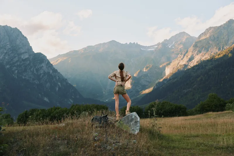 Young woman standing on a rock in a mountain valley, looking at distant peaks. Outdoor lifestyle concept with nature and adventure in warm daylight.