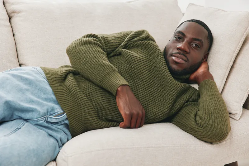 Relaxed Black man in a cozy olive sweater poses comfortably on a sofa, showcasing a casual and stylish indoor lifestyle with a serene expression
