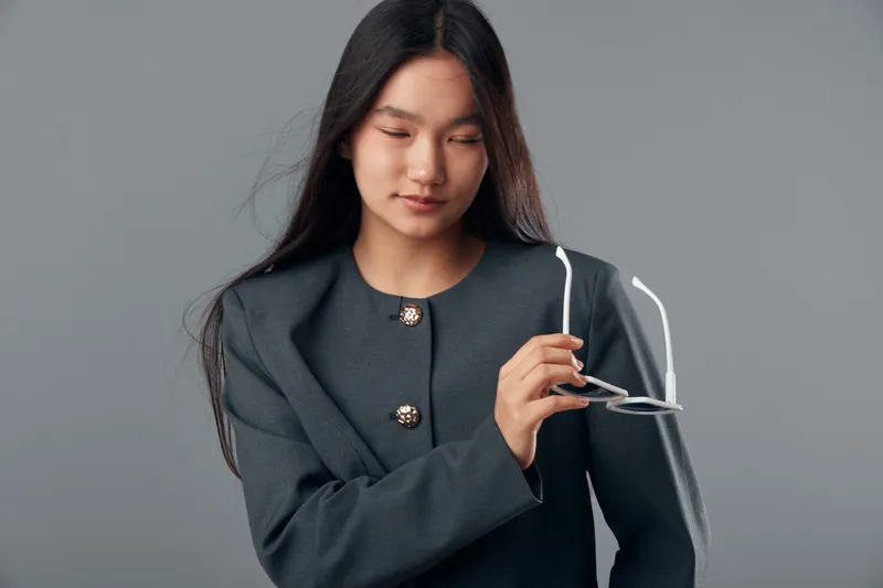 Young woman with long dark hair holding white eyeglasses and wearing gray jacket poses on gray background in studio. Female model is looking down with calm expression and straight hair.