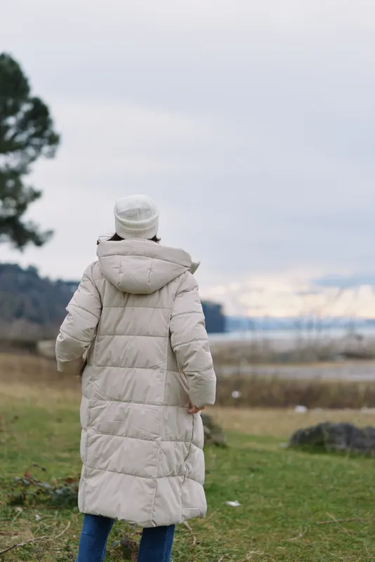 Person wearing a long beige winter coat stands in an open field with grass under a pale sky. The scene captures quiet solitude and seasonal fashion in a natural outdoor setting.