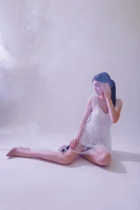 Young woman in a white lace dress sits on the floor with a soft dreamy expression. The minimalistic studio background adds elegance to this beauty and fashion concept.