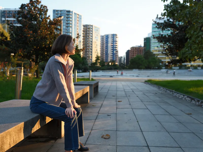 A thoughtful woman sits on a wooden bench in an urban park, enjoying sunlight. She gazes into the distance with a calm expression, surrounded by trees and modern buildings.