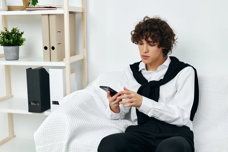 Кудрявый парень сидит на белом диване со смартфоном в руках