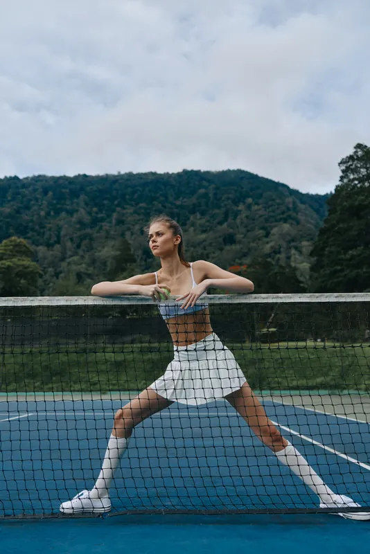 Stylish female tennis player in active wear striking a confident pose on a blue court with a mountainous backdrop, showcasing sports, fitness, and determination