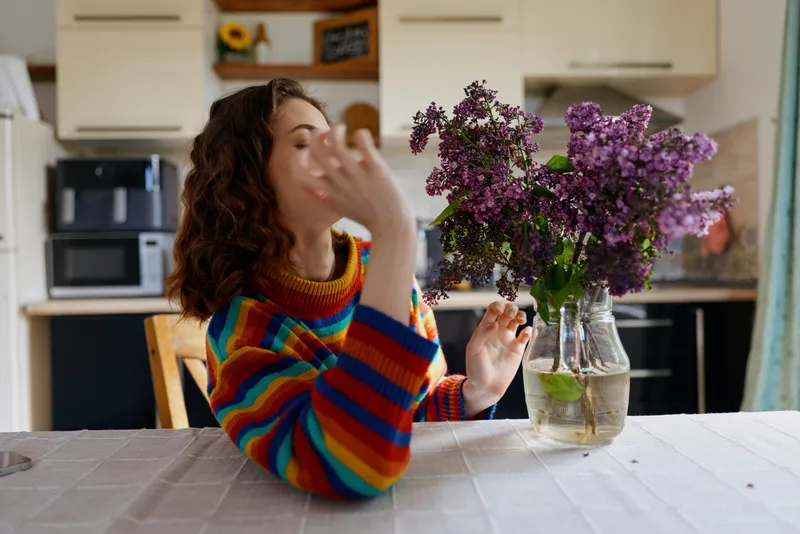 A young woman with curly hair in a colorful striped sweater sits at a bright kitchen table, reaching toward a glass vase of purple lilacs, capturing a casual everyday moment of calm concentration.