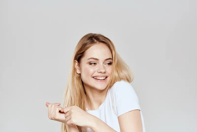 Милую блондинку в белой футболке, жестикулирующую руками, сняли в стиле лайфстайл на светлом фоне