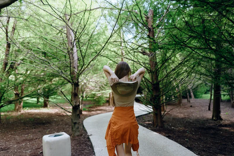 Woman walking on curved forest path between tall green trees, casual summer outfit and backpack, rear view of person enjoying nature, peaceful outdoor stroll in woodland.