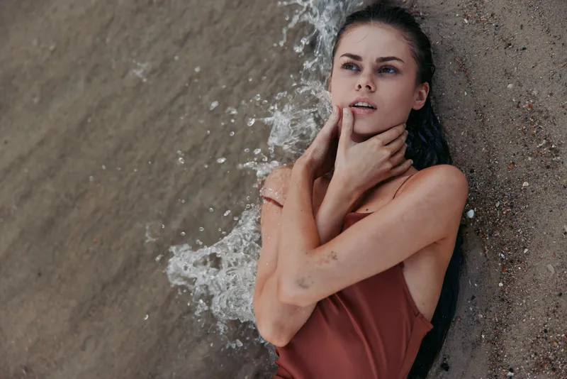 thoughtful woman lying on sandy shore in swimsuit with wet hair touching face, sea waves splashing, summer beach relaxation mood