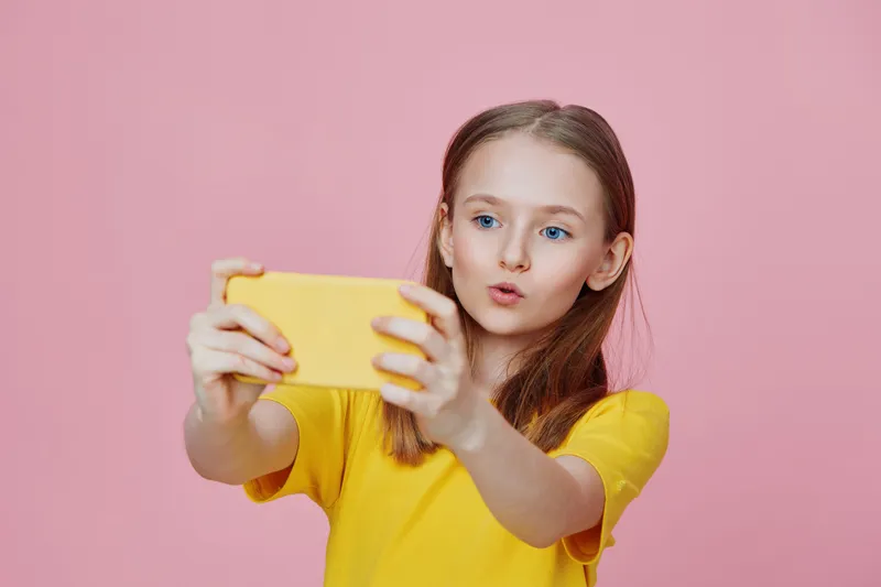 Young girl taking a selfie with a smartphone against a pink background, wearing a bright yellow shirt, showcasing happiness and confidence