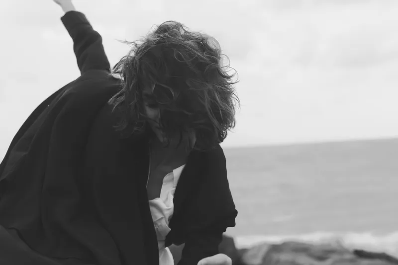 Gesture of a figure in wind by the sea, arm raised, moody black and white photography capturing motion and emotion