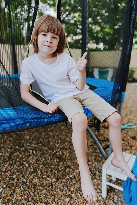 Cheerful boy giving a thumbs-up while sitting on a trampoline. The yard features gravel and greenery, creating an inviting outdoor atmosphere for play.