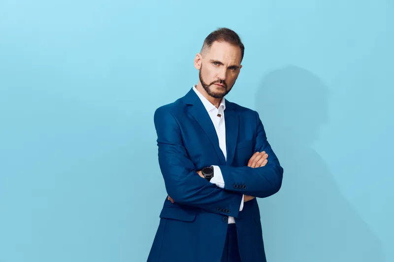 A professional businessman stands isolated against a blue background. The confident, composed figure conveys authority and steadiness, portraying ambition and focus in a modern corporate setting.