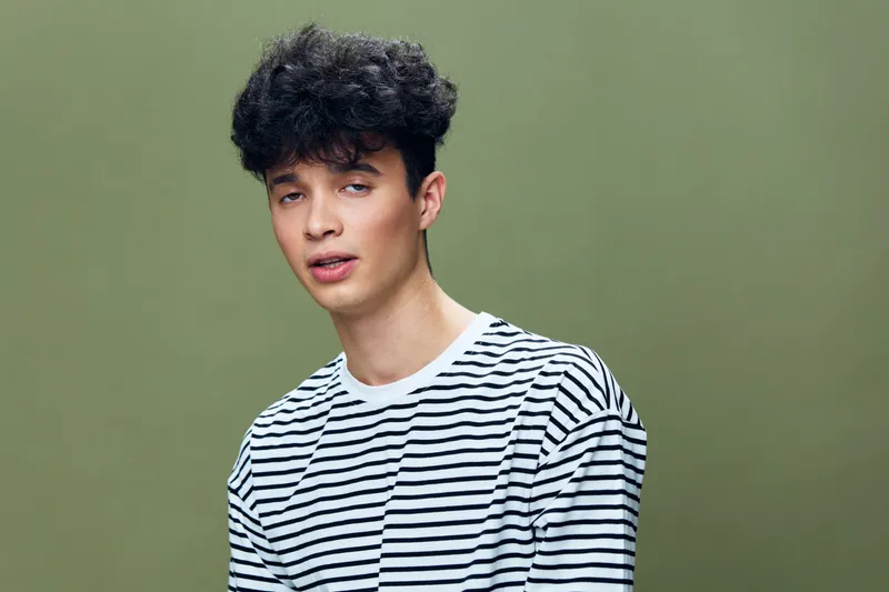 Young man with curly hair wearing a striped shirt against a green background, expressing a relaxed and casual mood in a simple studio setting.
