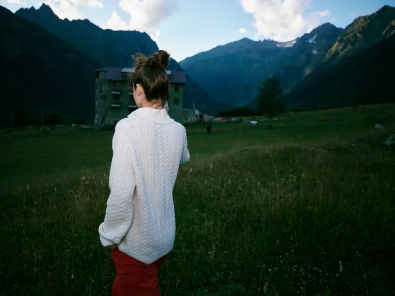 Back view of a woman in a white knitted sweater standing in a green field with distant mountains under a blue sky, conveying peaceful countryside and outdoor exploration