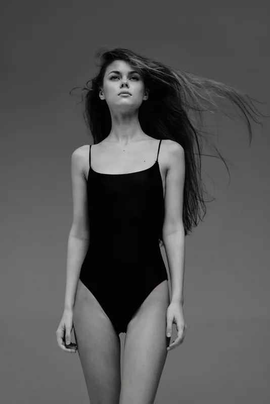 Black and white portrait of a young woman in a black swimsuit with flowing hair against a plain background.