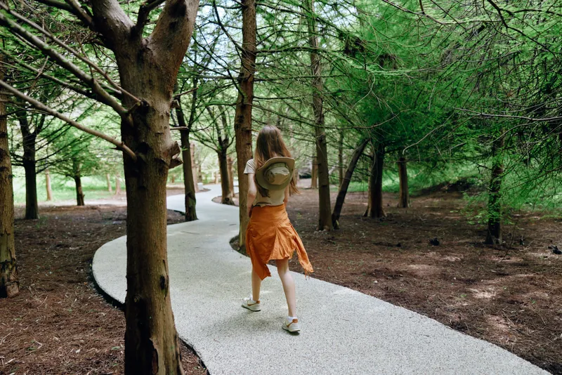 Woman with backpack walking along a curved path through forest and trees on a trail in nature, young hiker in orange skirt exploring outdoors, serene green scenery and peaceful journey