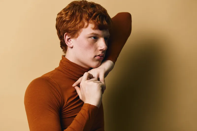 Redhead young man with curly hair wearing brown turtleneck posing with hand behind head and thoughtful expression in studio with beige background and soft lighting.