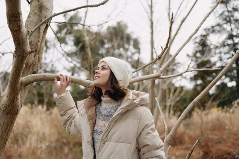 Woman in a warm coat and knit beanie gazes upward in a bare trees landscape, capturing a calm winter moment outdoors with soft natural light and muted colors.