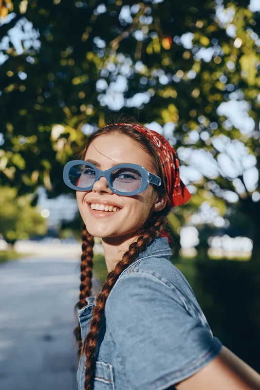 Lifestyle woman in modern boho-western denim street style smiling outdoors with film color effect, wearing blue sunglasses and red bandana under green tree shade.