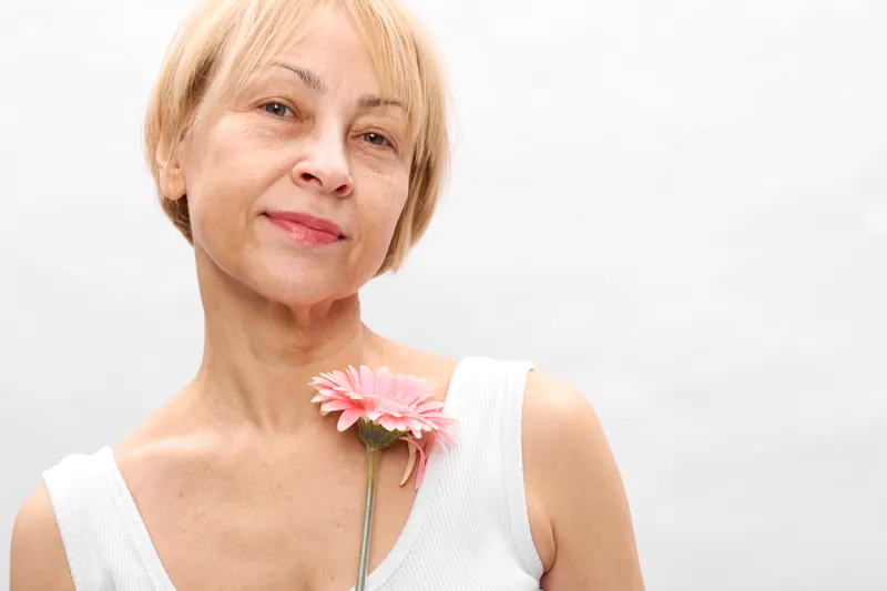 Woman portrait elderly blonde flower pink skincare wellness beauty white background, mature female holds gerbera, soft smile, calm wellness mood, studio light