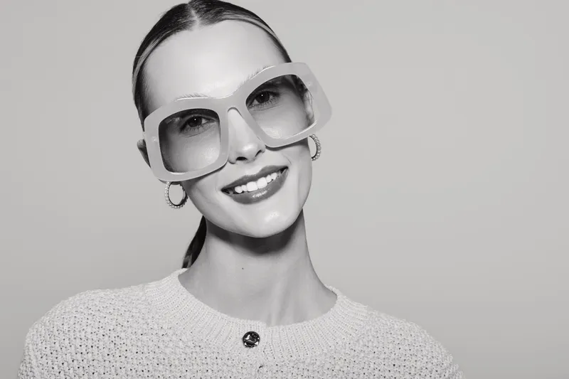 Smiling Caucasian woman with fair skin wearing oversized sunglasses and hoop earrings in studio. Concept of fashion, style, and confidence.