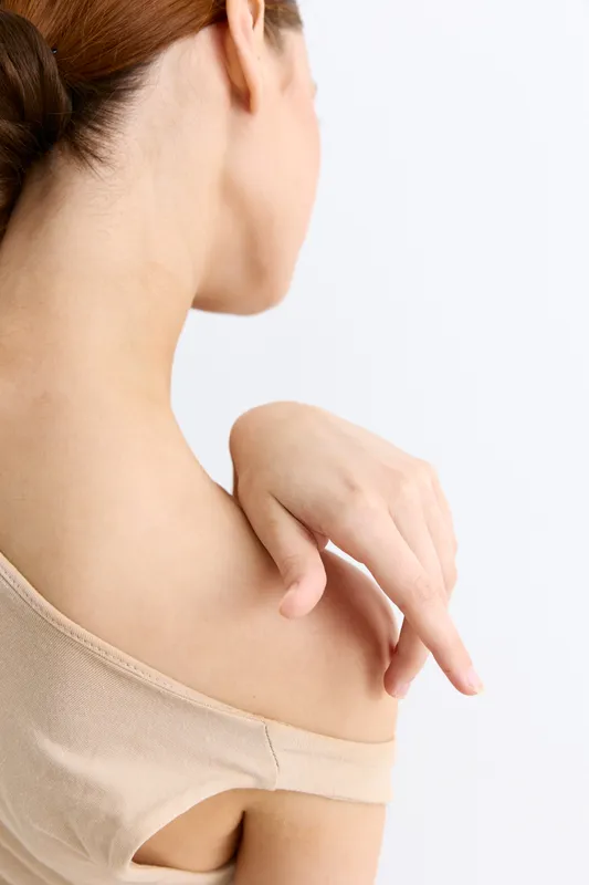 Woman touching her shoulder gently, showing skin care and relaxation with soft natural light and neutral background. Calm mood, minimalistic style.