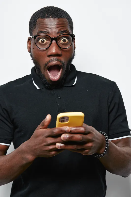 Black man with dark skin and glasses wearing black polo shirt looking surprised while holding yellow smartphone in hands isolated on white background with open mouth expression.