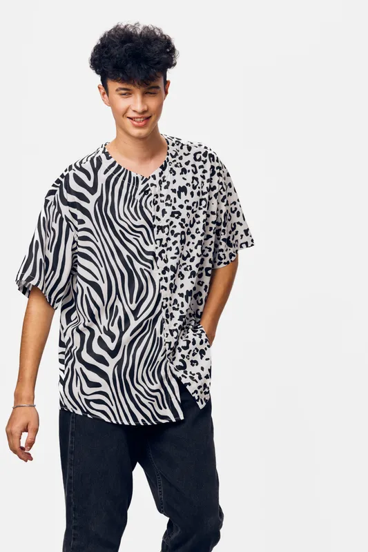 young man wearing a black and white animal print shirt with casual black pants posing with hands in pockets against a plain light background, smiling confidently