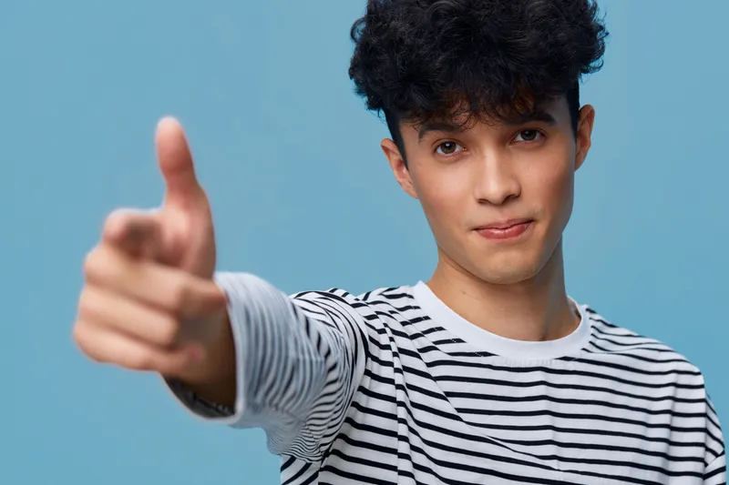 young man pointing finger towards camera with confident expression wearing striped shirt against blue background, casual style portrait