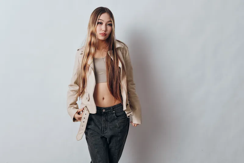 Young woman standing against plain light background with long hair wearing casual beige jacket and crop top paired with black jeans in studio portrait fashion style.