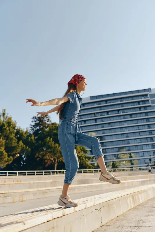 lifestyle woman in modern boho-western denim street style jumps outdoors with clear blue sky and urban building, vibrant film color fashion and casual vibe