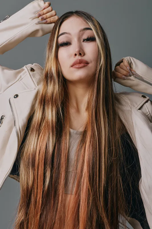 Young woman with long straight hair wearing light jacket posing with arms raised studio portrait. Fashion model with natural makeup and neutral expression on grey background.